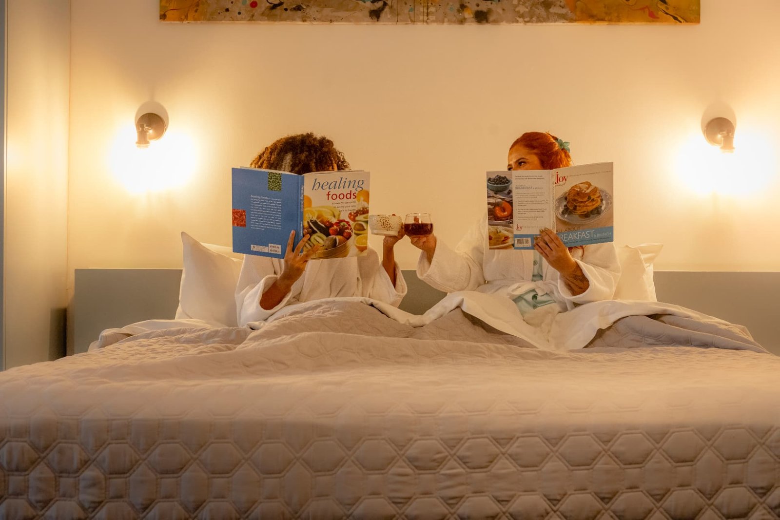 Dos mujeres en bata leyendo y brindando en la cama de una habitación de Tántalo